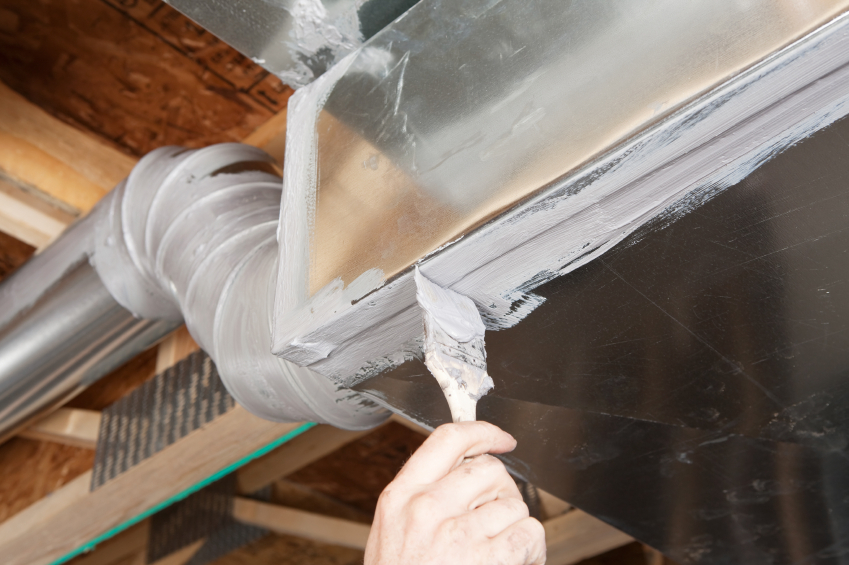 Hand applying gray sealant to an HVAC duct joint with a putty knife.