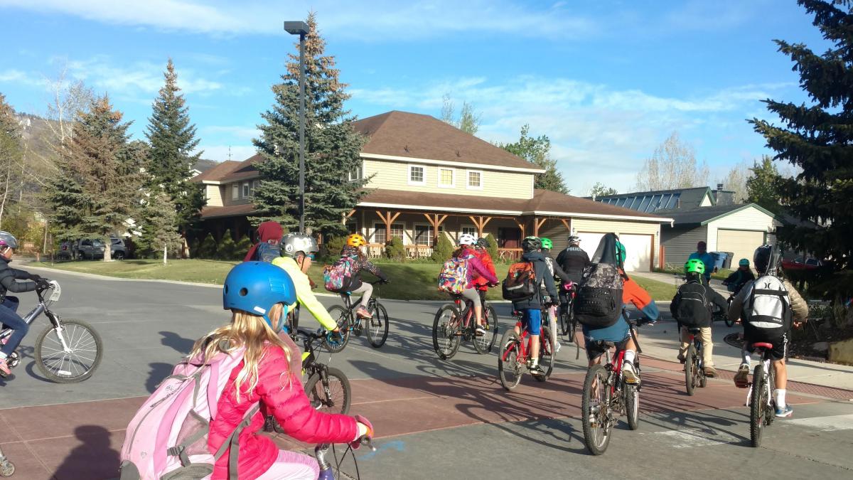 Group of children and adults on bicycles waiting at a suburb intersection with a stop sign, residential houses, and coniferous trees in the background under a clear blue sky.